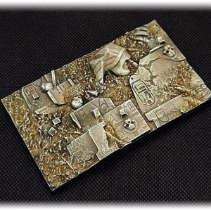 May include: A rectangular, miniature diorama base with a detailed, weathered appearance. The base features a textured, tan and brown surface with sculpted ruins, skulls, and hieroglyphs, suggesting an ancient, desert-themed setting. The color palette is primarily muted, with shades of gray and brown.