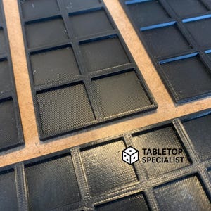 Op de afbeelding: Close-up van verschillende zwarte, rechthoekige speltrays met een rasterpatroon. De trays zijn gemaakt van een textuurrijk materiaal en zijn ontworpen om spelstukken in te bewaren. Het logo "TABLETOP SPECIALIST" is zichtbaar.
