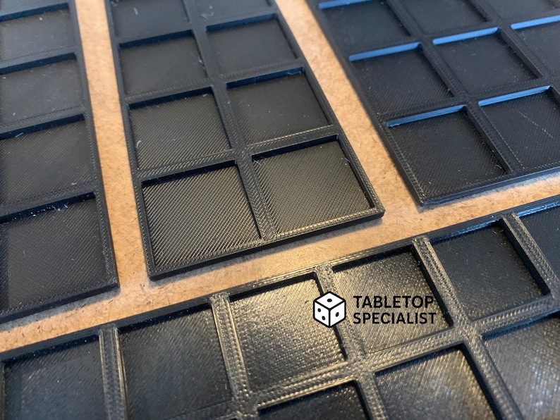 Op de afbeelding: Close-up van verschillende zwarte, rechthoekige speltrays met een rasterpatroon. De trays zijn gemaakt van een textuurrijk materiaal en zijn ontworpen om spelstukken in te bewaren. Het logo "TABLETOP SPECIALIST" is zichtbaar.
