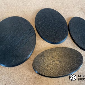 Op de afbeelding: Vier zwarte, ovaalvormige spelbases van verschillende afmetingen. De bases hebben een textuuroppervlak en zijn gerangschikt op een lichtbruin oppervlak. De bases zijn waarschijnlijk voor miniatuurfiguren die in tabletopspellen worden gebruikt. Het logo "TABLETOP SPECIALIST" staat rechtsonder.