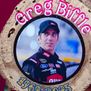 May include: A wooden plaque with a photo of Greg Biffle, framed in a circular design. The name "Greg Biffle" is in pink letters. The years "1969-2025" are in blue. The photo shows Biffle in a racing uniform.