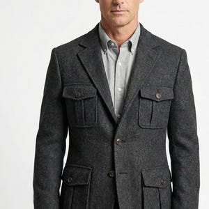 Op de afbeelding: Een antracietgrijze blazer met vier voorzakken en knoopsluitingen. De jas wordt gedragen over een lichtgrijs button-down overhemd. De tekst "THA FASHION TAILOR" is linksboven zichtbaar. De blazer is gemaakt van een textuurstof.