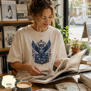 Può includere: T-shirt color avorio con una grafica dettagliata di un gufo blu con le ali spiegate, appollaiato su funghi blu. Il gufo ha gli occhi rossi ed è circondato da un design decorativo. La persona sta leggendo un libro.