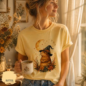 Può includere: T-shirt color burro con un'illustrazione di una marmotta con un cappello da strega, sullo sfondo di una luna crescente e stelle. Il disegno è circondato da foglie autunnali. La maglietta ha le maniche arrotolate.