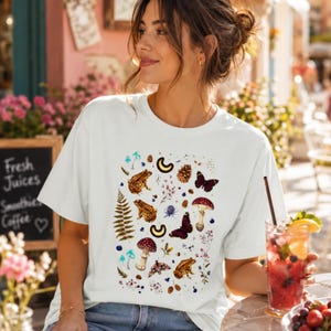 Può includere: T-shirt bianco con un design a tema natura. La grafica include illustrazioni di rane marroni, funghi con cappello rosso, farfalle e altri elementi botanici. La maglietta è uno stile casual e confortevole.