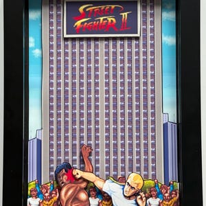 May include: Framed artwork featuring a scene from Street Fighter II. The image depicts two characters in combat with a cityscape backdrop and the game's logo. The artwork is encased in a black frame.