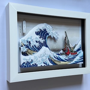 Pode incluir: Obra de arte emoldurada retratando uma onda oce&acirc;nica estilizada com barcos. A onda &eacute; renderizada em tons de azul e branco, com barcos amarelos e vermelhos. A obra de arte est&aacute; em uma moldura branca.