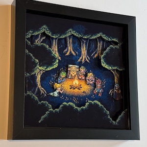 Puede incluir: Una obra de arte en caja de sombra enmarcada que representa una escena de fogata pixelada en un bosque. La obra presenta varios personajes reunidos alrededor de un fuego, con árboles y follaje en el fondo. El marco es negro.