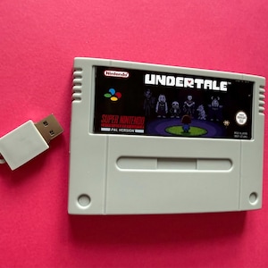 May include: A light grey Super Nintendo game cartridge featuring an Undertale label with pixelated characters and the text UNDERTALE, PAL VERSION, and Nintendo. Next to the cartridge is a small, rectangular white USB flash drive with a metal connector.