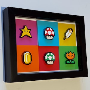 May include: Framed artwork with pixelated video game icons. The icons include a yellow star, a red and white mushroom, a white feather, a gold coin, a green and white mushroom, and an orange flower. The icons are set against colourful backgrounds.