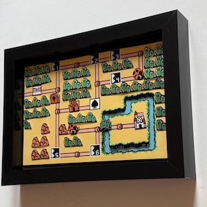 Könnte beinhalten: Ein 3D-Schattenbox-Rahmen mit einer mehrschichtigen Papierkunst-Nachbildung einer Retro-Videospiel-Weltkarte. Die gelbe Karte zeigt grüne pixelige Büsche, orangefarbene Felsformationen, nummerierte Level-Markierungen, ein Schloss und einen Startpunkt.