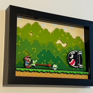 Op de afbeelding: Een ingelijste 3D pixel art scène met Mario op Yoshi, een groen schildpadachtig wezen, met een rode tong uitgestrekt naar een groene paddenstoel. Een zwart-witte Bullet Bill staat op de achtergrond. De scène speelt zich af tegen een groene en bruine achtergrond.