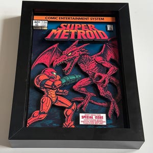 May include: Framed artwork with a 3D layered design of a Super Metroid comic book cover. The artwork shows a red armored character fighting a red dragon-like creature. The frame is black, and the artwork includes the text "COMIC ENTERTAINMENT SYSTEM".