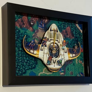 Puede incluir: Obra de arte enmarcada que presenta una nave espacial blanca y dorada con personajes pixelados, sobre un fondo de paisaje detallado. La obra de arte está encerrada en un marco negro, creando un efecto 3D.