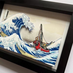 Pode incluir: Obra de arte emoldurada retratando um barco vermelho com um personagem navegando por uma grande onda oce&acirc;nica. A onda &eacute; azul e branca, ao estilo da arte tradicional japonesa. A moldura &eacute; preta.