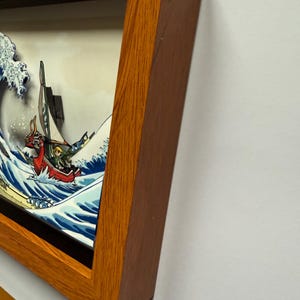 Pode incluir: Uma obra de arte emoldurada que retrata um barco estilizado navegando em uma grande onda, com um personagem no barco. A obra de arte est&aacute; em uma moldura de madeira. A onda &eacute; azul e branca, e o barco &eacute; vermelho.