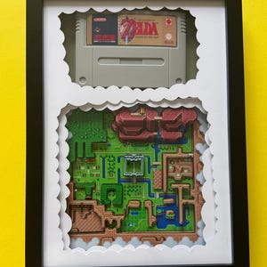 May include: A black framed shadow box containing a gray Super Nintendo game cartridge for The Legend of Zelda: A Link to the Past, positioned above a multi-layered 3D paper diorama depicting the game's overworld map with green and brown terrain.