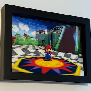 Puede incluir: Una obra de arte 3D enmarcada que representa una escena de videojuego. La obra muestra un personaje con gorro rojo y peto azul sobre una plataforma colorida. El fondo muestra un edificio con escaleras y un cielo azul.