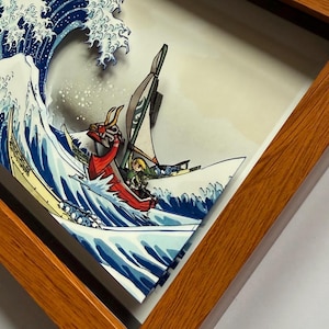 Pode incluir: Obra de arte emoldurada retratando uma cena de ondas japonesas estilizadas com um barco. O barco apresenta uma figura de proa de drag&atilde;o vermelho e um personagem de videogame. A obra de arte est&aacute; em uma moldura de madeira.