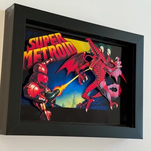 May include: Framed artwork depicting the Super Metroid video game. A red character fires a beam at a red dragon-like creature. The words "SUPER METROID" are in yellow and red. The artwork is presented in a black frame.