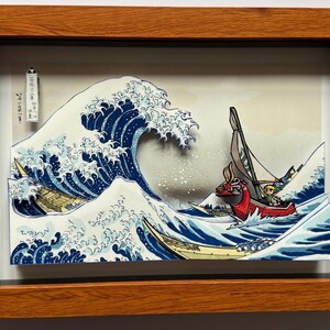 Pode incluir: Obra de arte 3D emoldurada retratando uma onda oce&acirc;nica estilizada com barcos, inspirada na arte japonesa. A onda &eacute; azul escuro com espuma branca, e os barcos s&atilde;o vermelhos e amarelos. A obra de arte est&aacute; em uma moldura de madeira.