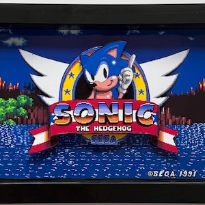 May include: Framed artwork featuring Sonic the Hedgehog, the iconic video game character. The image shows Sonic with a thumbs-up, set against a pixelated background of a blue sky, water, and land. The artwork includes the game's logo and the SEGA copyright.