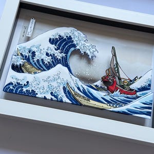 Pode incluir: Uma obra de arte 3D emoldurada que retrata uma onda oce&acirc;nica estilizada em tons de azul e branco, com um barco vermelho e uma figura navegando pelas ondas. A obra de arte &eacute; emoldurada em uma moldura branca.