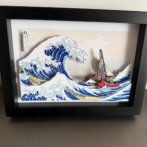 Pode incluir: Obra de arte 3D emoldurada que retrata uma onda oce&acirc;nica estilizada com um barco. A onda &eacute; renderizada em tons de azul e branco, com um barco vermelho e amarelo. A obra de arte est&aacute; em uma moldura preta.