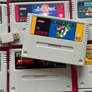 May include: A grey Super Nintendo video game cartridge featuring a Super Mario World label with Mario riding Yoshi. The cartridge has a yellow top section, a black Nintendo logo, and a blue background. A small white USB drive is positioned to the left.