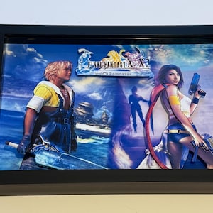 May include: Framed artwork depicting characters from Final Fantasy X/X-2. The image shows a male character with a sword and a female character holding a gun, set against a blue and white background. The title "Final Fantasy X/X-2 HD Remaster" is displayed.