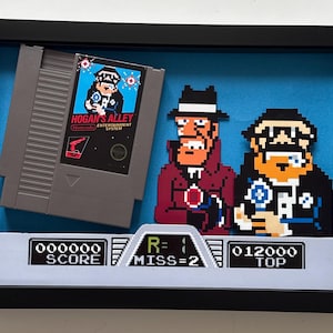 Może przedstawiać: Oprawiona w ramkę trójwymiarowa ekspozycja zawierająca szarą kasetę z grą Hogan's Alley na konsolę Nintendo Entertainment System, dwie pikselowe wycinanki postaci oraz panel interfejsu gry z tekstem SCORE 000000, R=1, MISS=2 i TOP 012000.
