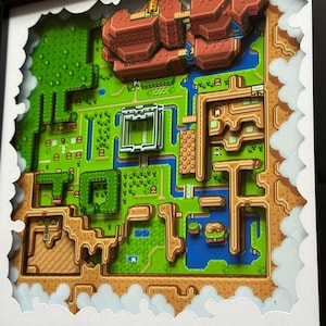 May include: A framed paper art piece showing a layered, pixelated map of a video game world. The artwork features a green landscape with brown mountains, blue water, and structures. The frame is black, and the edges are cut to resemble clouds.