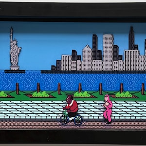 May include: Framed 3D artwork showing a pixelated New York City scene. The artwork features the Statue of Liberty, a cityscape, and figures on a sidewalk. A person rides a bicycle, and another walks. The background is a blue sky.