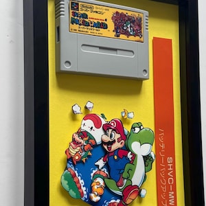 May include: A framed Super Mario World Super Famicom game cartridge mounted above a 3D paper cutout display featuring Mario, Yoshi, and Bowser on a bright yellow background. The vertical orange label includes the text SHVC-MW and Japanese characters.
