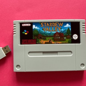 Puede incluir: Un cartucho de videojuego gris de Super Nintendo para Stardew Valley con una colorida ilustración de paisaje en la etiqueta, acompañado de una pequeña unidad flash USB blanca con un puerto azul, sobre un fondo rosa sólido.