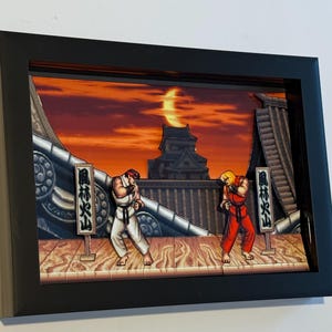 May include: Framed 3D artwork featuring a fighting game scene with two characters in a dojo setting. One character is in white, the other in red. The background shows a sunset and Japanese text.