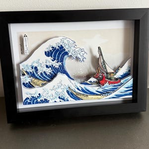Pode incluir: Uma obra de arte em papel 3D emoldurada que retrata uma onda oce&acirc;nica estilizada com barcos. A obra de arte tem uma moldura preta, com a onda renderizada em tons de azul e branco. Um barco vermelho com uma vela verde est&aacute; no centro.