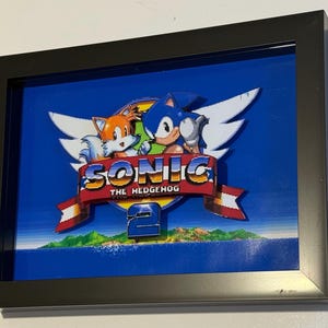 May include: Framed artwork featuring the Sonic the Hedgehog 2 logo. The image shows Sonic and Tails with the game title on a red banner. The background is blue with pixelated details, all encased in a black frame.