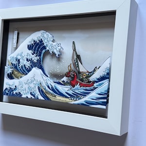 Pode incluir: Obra de arte emoldurada retratando uma cena de ondas estilizada com um barco. O barco apresenta uma figura de proa de drag&atilde;o vermelho e um personagem com uma t&uacute;nica verde. A obra de arte est&aacute; em uma moldura branca com uma borda preta.