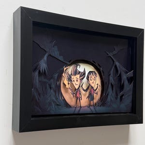 May include: A layered shadow box art piece in a black frame featuring two cartoon characters standing before a large, glowing circular moon. The scene includes dark, silhouetted trees and foliage surrounding the figures, who are holding small torches.