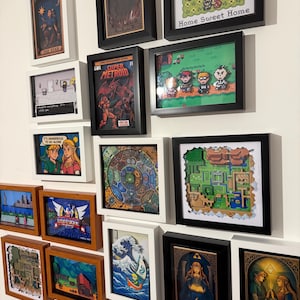 Puede incluir: Una colección de arte en cajas de sombra enmarcadas que presentan varias escenas y personajes de videojuegos. Las piezas incluyen pixel art, ilustraciones estilo tarot y diseños de mapas en marcos negros, blancos y de madera.