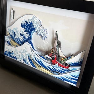 Pode incluir: Uma obra de arte em papel 3D emoldurada que retrata uma onda oce&acirc;nica estilizada com um barco. A onda &eacute; azul e branca, com um barco vermelho e verde. A obra de arte est&aacute; em uma moldura preta.