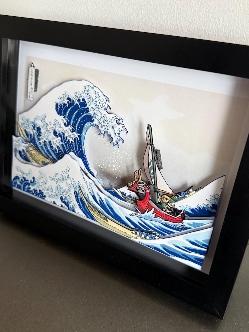 Pode incluir: Uma obra de arte em papel 3D emoldurada que retrata uma onda oce&acirc;nica estilizada com um barco. A onda &eacute; azul e branca, com um barco vermelho e verde. A obra de arte est&aacute; em uma moldura preta.