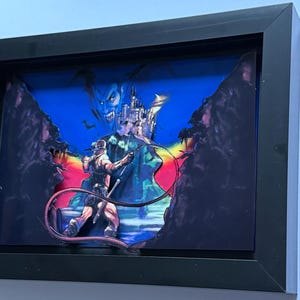以下が含まれることがあります： ビデオゲームのシーンを描いた額入りのアートワーク。キャラクターが城と不気味な姿に対してムチを振るっています。アートワークは、鮮やかな色とレイヤー効果があり、濃い青色の背景に設定され、黒いフレームに収められています。