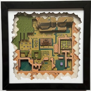 Op de afbeelding: Een ingelijst, gelaagd papieren kunstwerk dat een gedetailleerde kaart van een videogame-wereld weergeeft. De kaart toont verschillende terreinen, waaronder bergen, bossen en waterpartijen, in tinten groen, bruin en beige. Het kunstwerk is ingekaderd in een zwart frame.