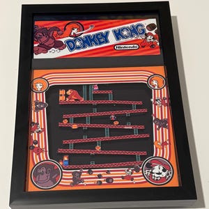 Könnte beinhalten: Gerahmtes Donkey Kong-Kunstwerk mit dem Logo des Spiels und einer Szene aus dem klassischen Arcade-Spiel. Das Kunstwerk hat einen schwarzen Rahmen und ein leuchtendes Orange- und Rot-Farbschema. Das Nintendo-Logo ist sichtbar.
