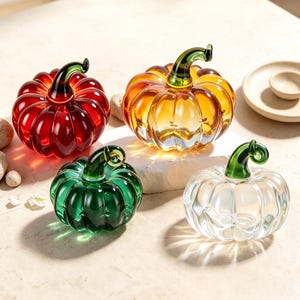 May include: Four glass pumpkins in red, orange, green, and clear, each featuring a distinct ribbed texture and a smooth, curved green stem. The translucent glass objects are arranged in a cluster, reflecting light to create bright highlights.