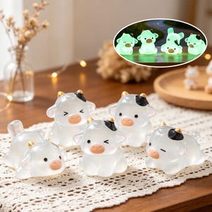 May include: A collection of five small, translucent cow figurines made of smooth material. Each cow features a white body, a black patch on the head, gold-coloured horns, and a pink snout. An inset shows the figurines glowing with a soft green light.