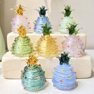 May include: A collection of eight small, handcrafted glass pineapple figurines in various pastel colors including blue, green, yellow, and pink. Each piece features a coiled glass body with small raised dots and a distinct green or yellow glass top.
