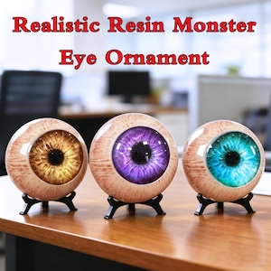 May include: Three spherical resin monster eye ornaments on black stands. Each features a realistic sclera with red veins and a distinct iris color: gold, purple, and teal. The text "Realistic Resin Monster Eye Ornament" is displayed above.